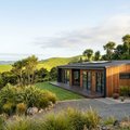 Single-storey timber-clad home with rooftop solar panels and a wooden deck among cabbage trees and rolling green hills